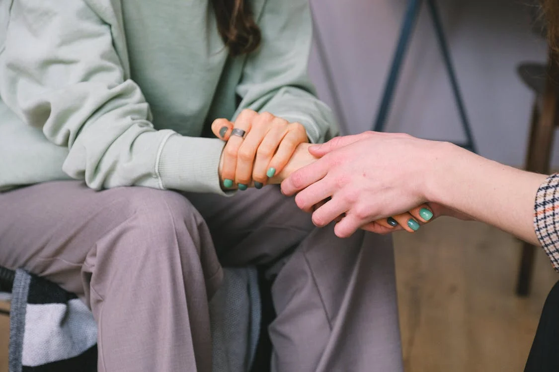 Close-up of two people's hands clasped together in a supportive gesture, one wearing a green sweater and the other with teal nail polish.