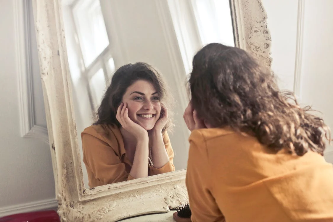 A smiling young woman with dark hair looking at herself in an ornate mirror, wearing a yellow top.
