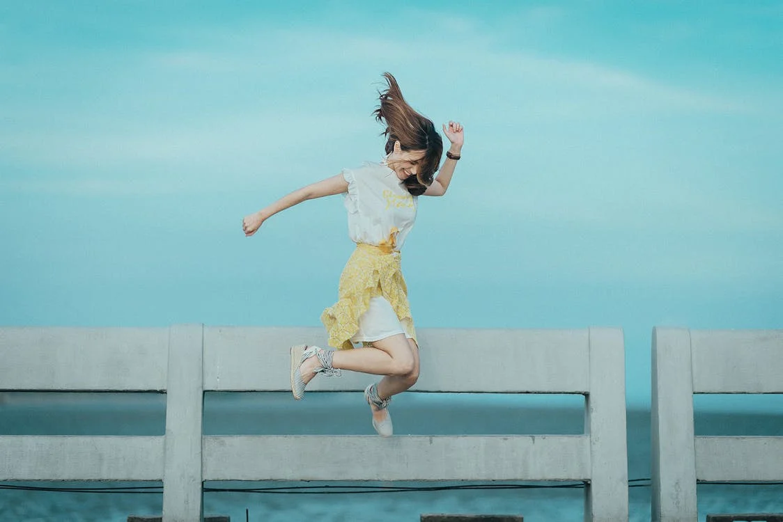 A woman in a white top and yellow skirt jumping joyfully on a concrete barrier against a bright blue sky.
