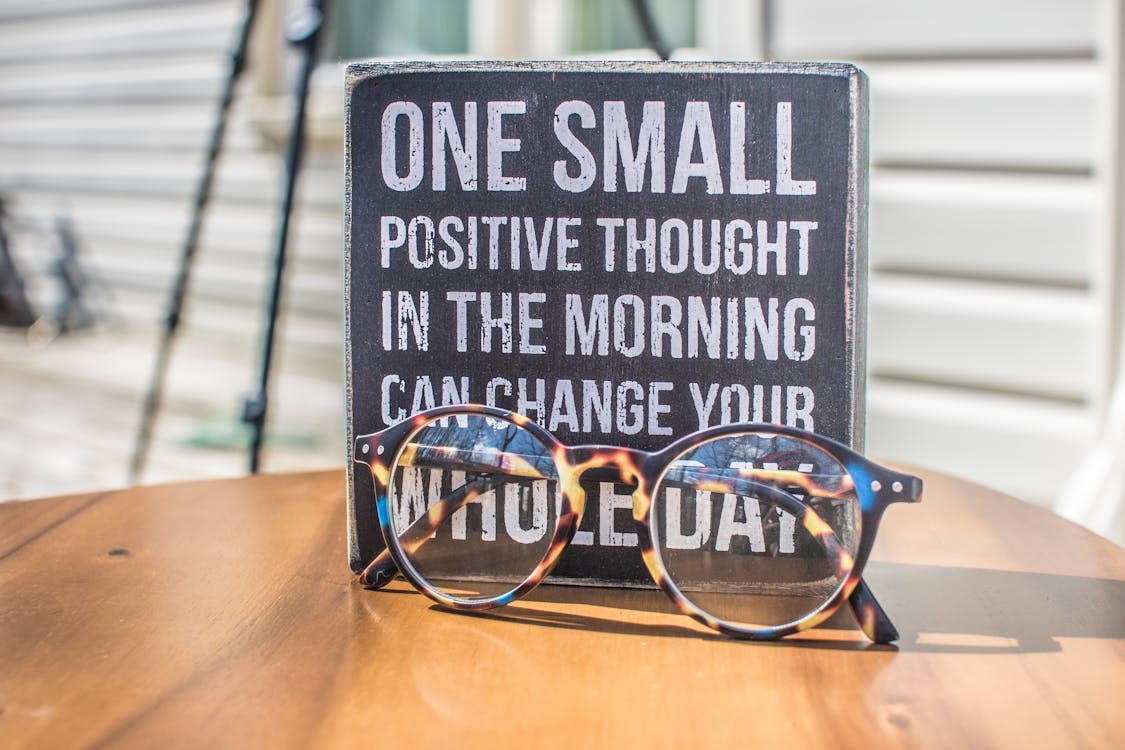 A wooden sign with the message "One small positive thought in the morning can change your whole day" and a pair of glasses in front.