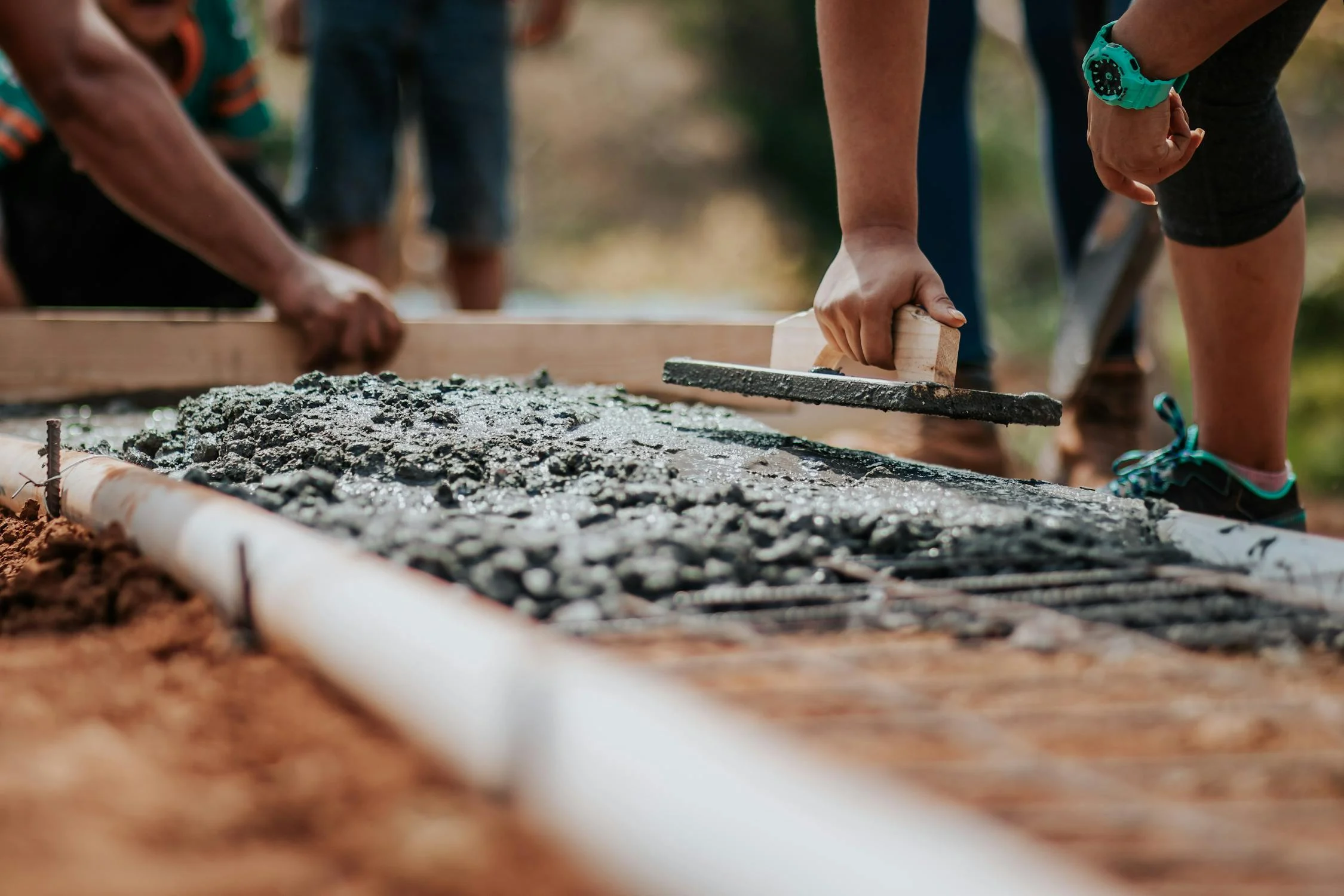 Close-up of hands smoothing wet concrete with tools, showing people working together on a construction project outdoors with one person wearing a turquoise watch.