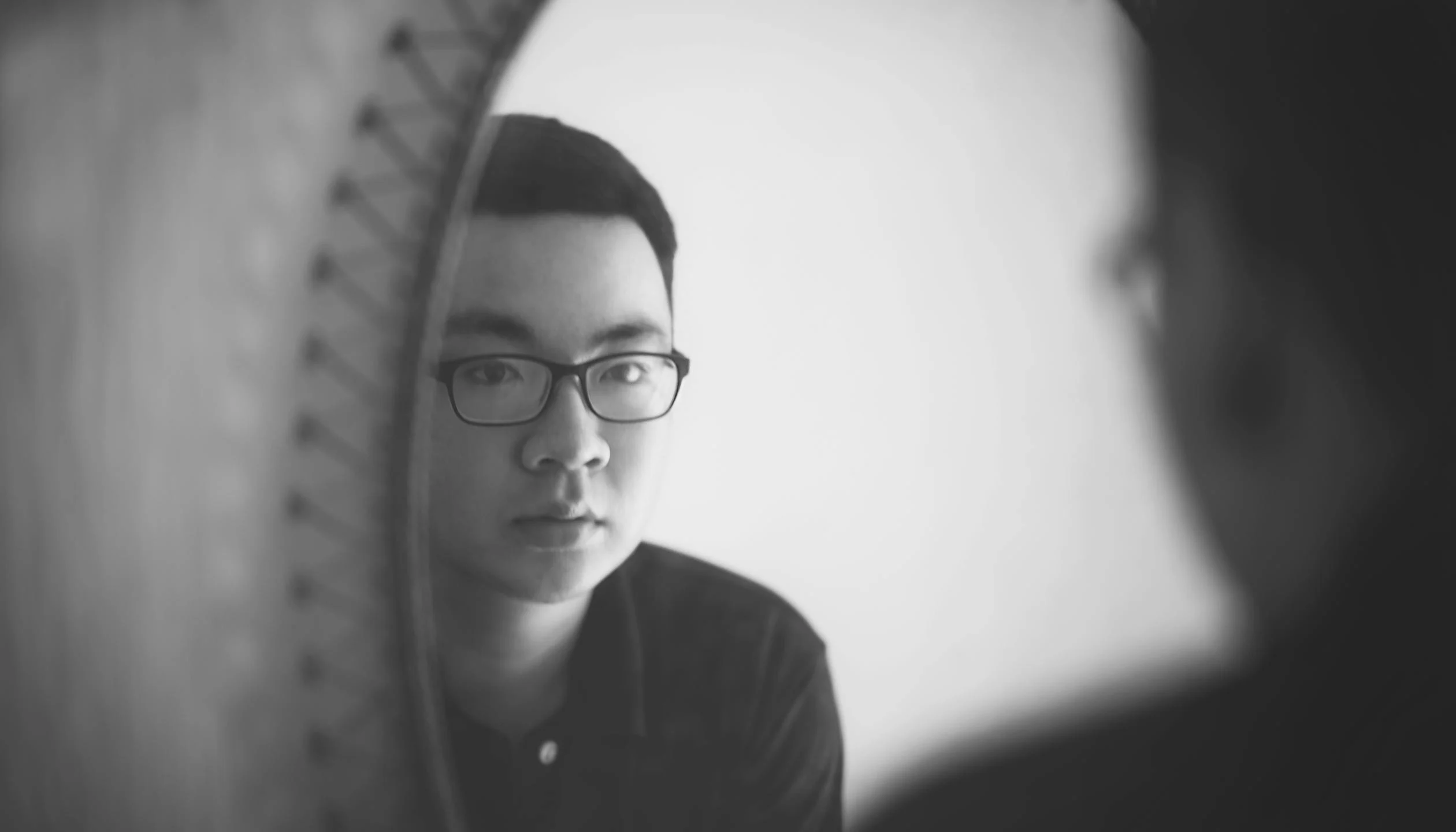 Black and white photograph showing a person with glasses looking thoughtfully in a mirror, creating a reflective, introspective mood with soft focus elements