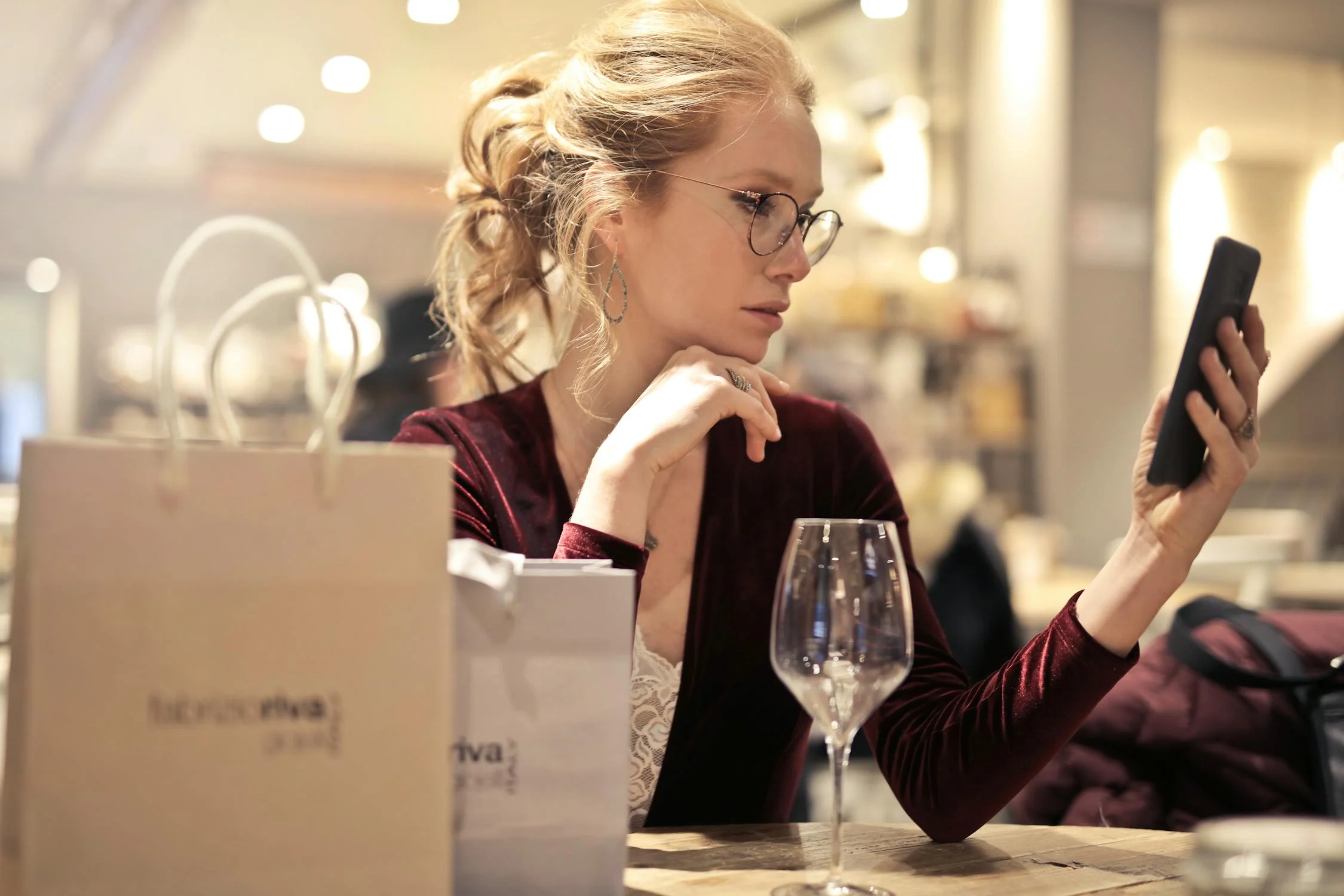 Woman with glasses and blonde hair in a burgundy velvet top intently looking at her smartphone while sitting at a restaurant table with a wine glass and shopping bags nearby.